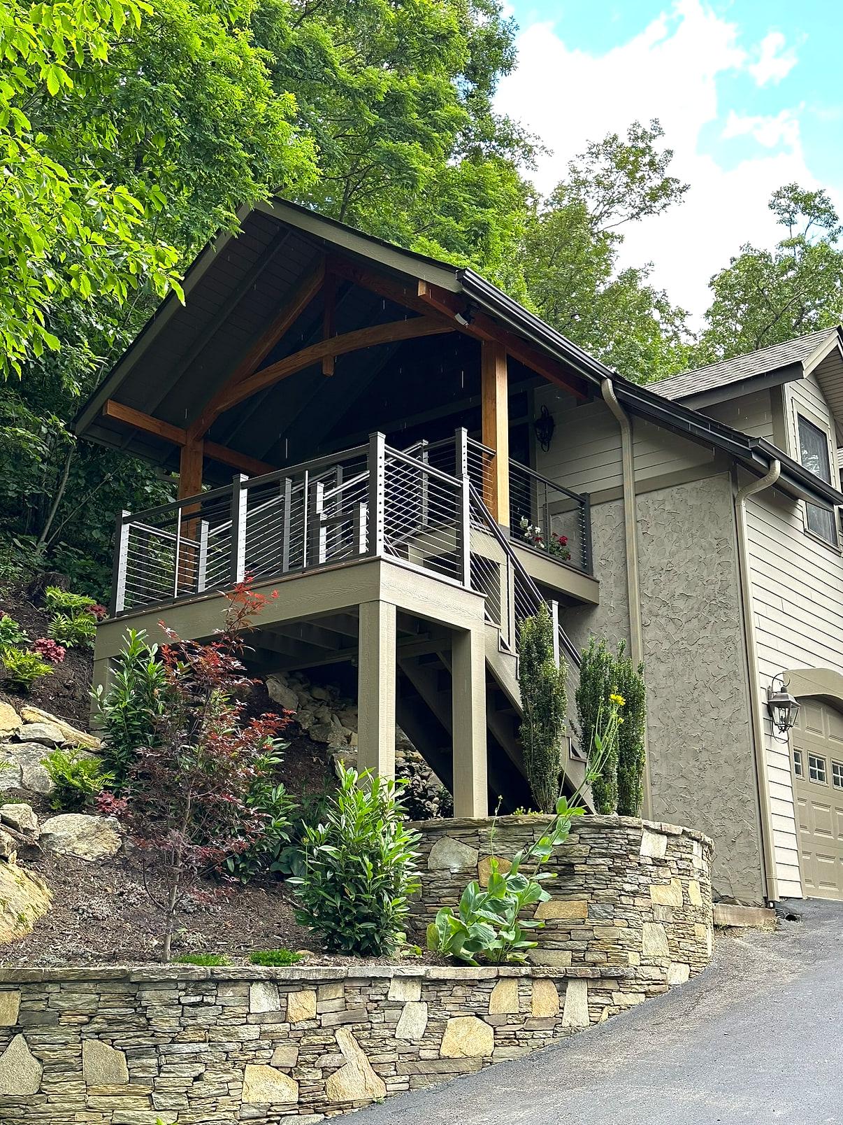 The same addition finished — timber-framed gable porch, cable railing, painted stair and stacked-stone terraces planted out