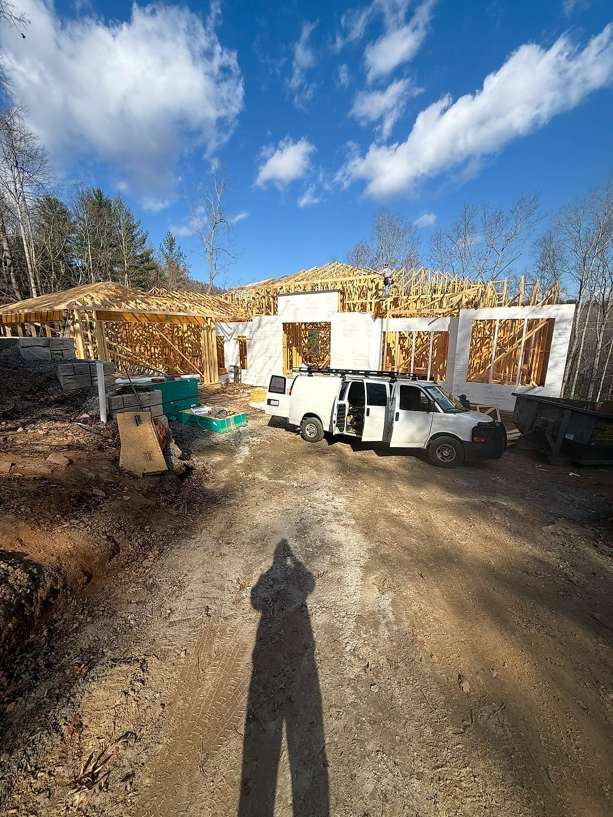 New-construction shell framed out — roof trusses being set over house-wrapped walls, with the crew van parked on the graded drive